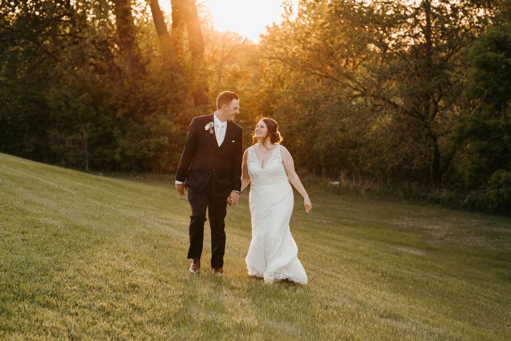 Happy bride and groom walking hand in hand on grass during warm golden hour sunset.