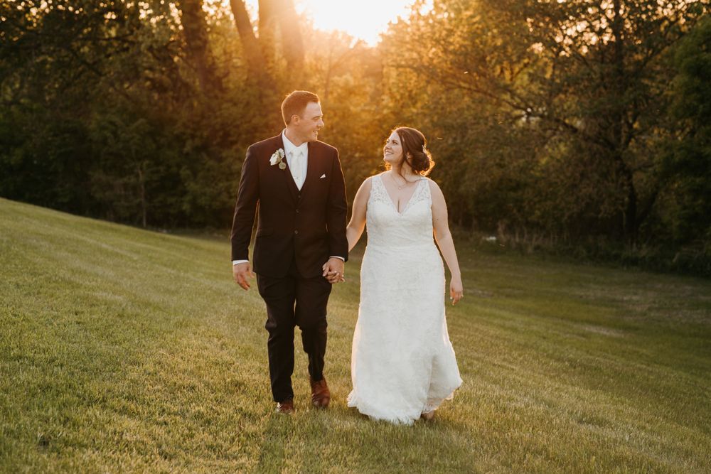 Smiling bride and groom holding hands while strolling across lawn in glowing sunset light.