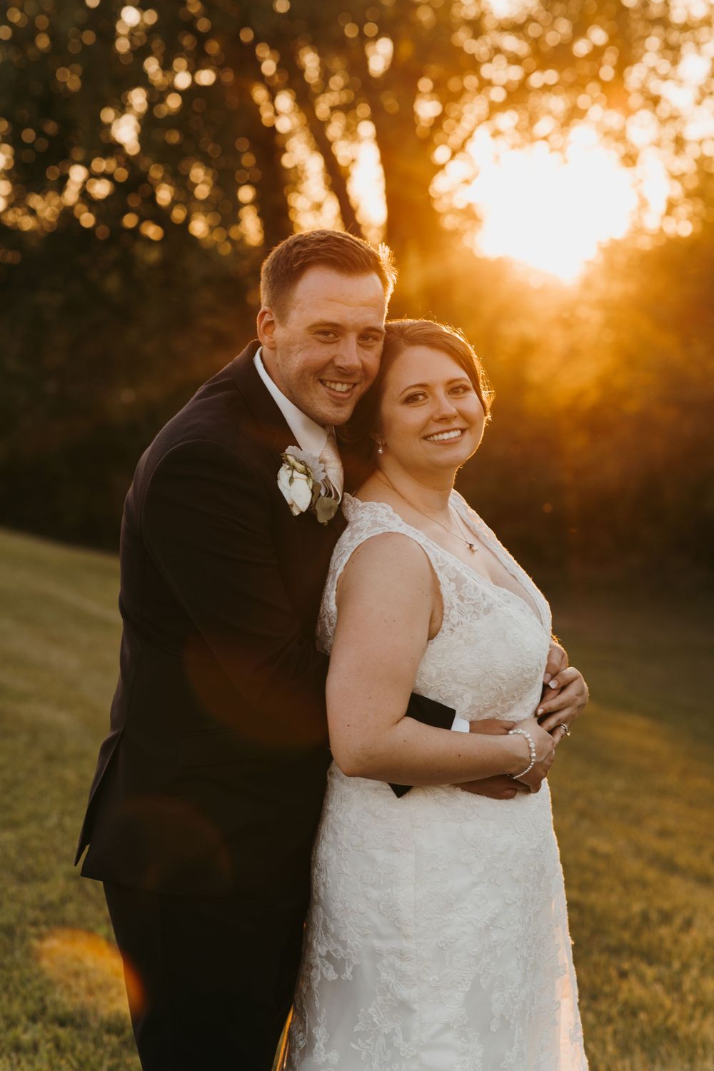 Groom embraces bride from behind as they smile together during a glowing golden hour wedding portrait.