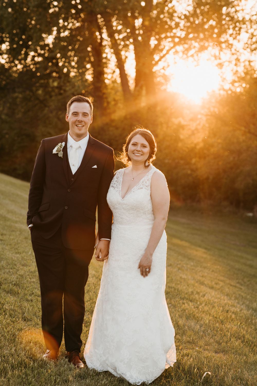 Bride and groom hold hands and smile during a radiant golden hour outdoor wedding portrait.