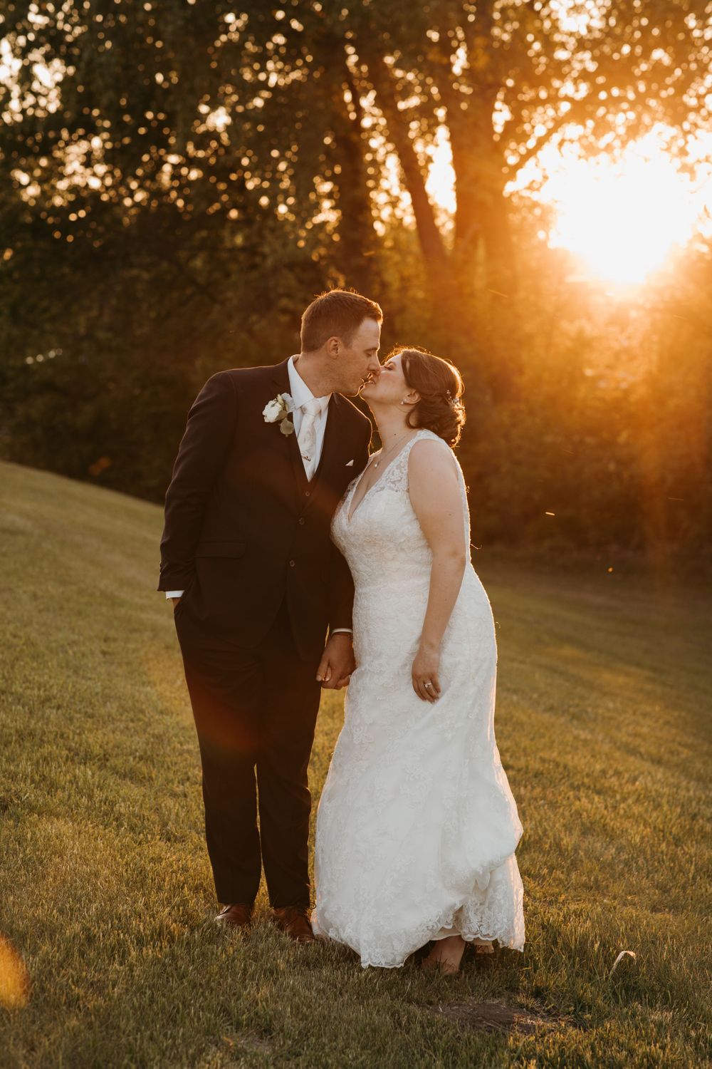 Bride and groom share a romantic sunset kiss on a grassy hillside during their outdoor wedding.
