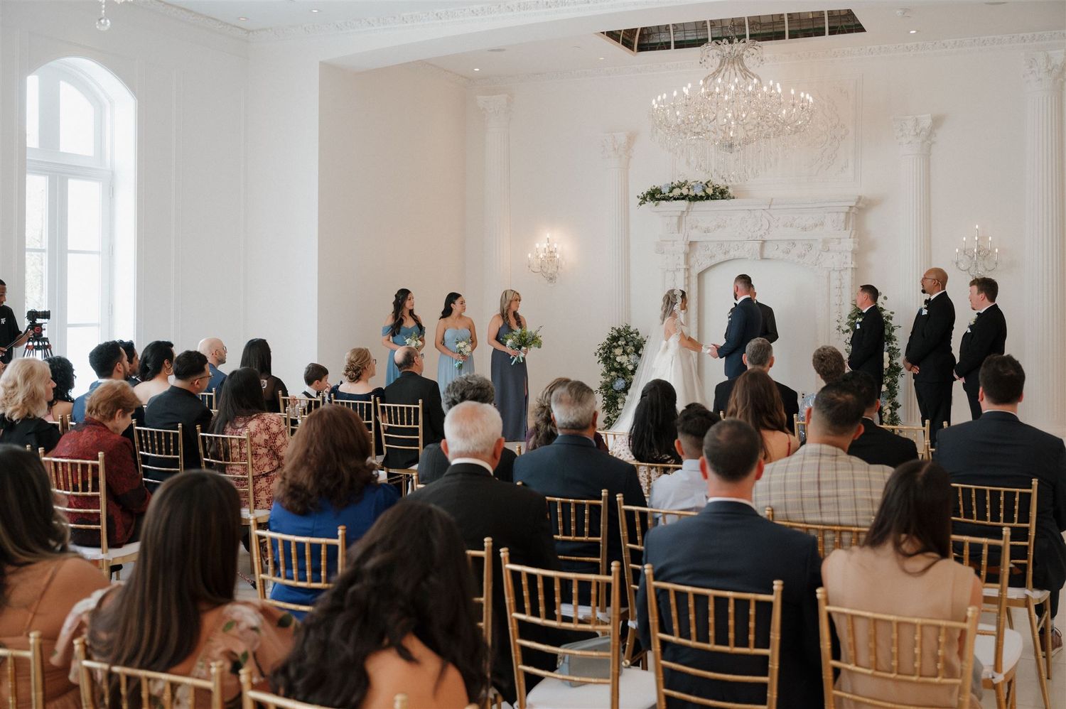 Wedding guests seated in gold chairs watch a ceremony in an elegant white room with chandeliers.