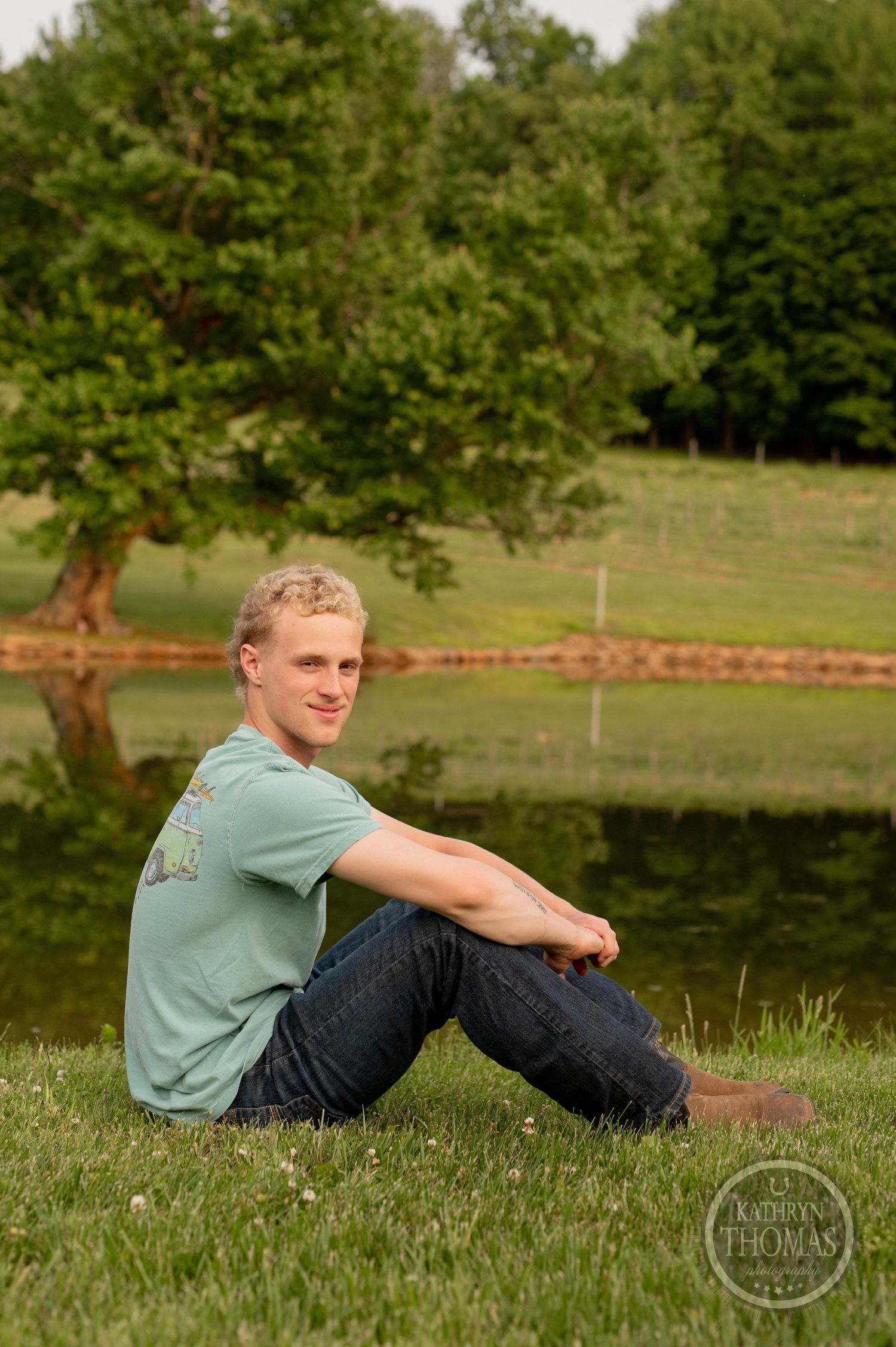 Person in casual attire sits on grass overlooking tranquil pond landscape.