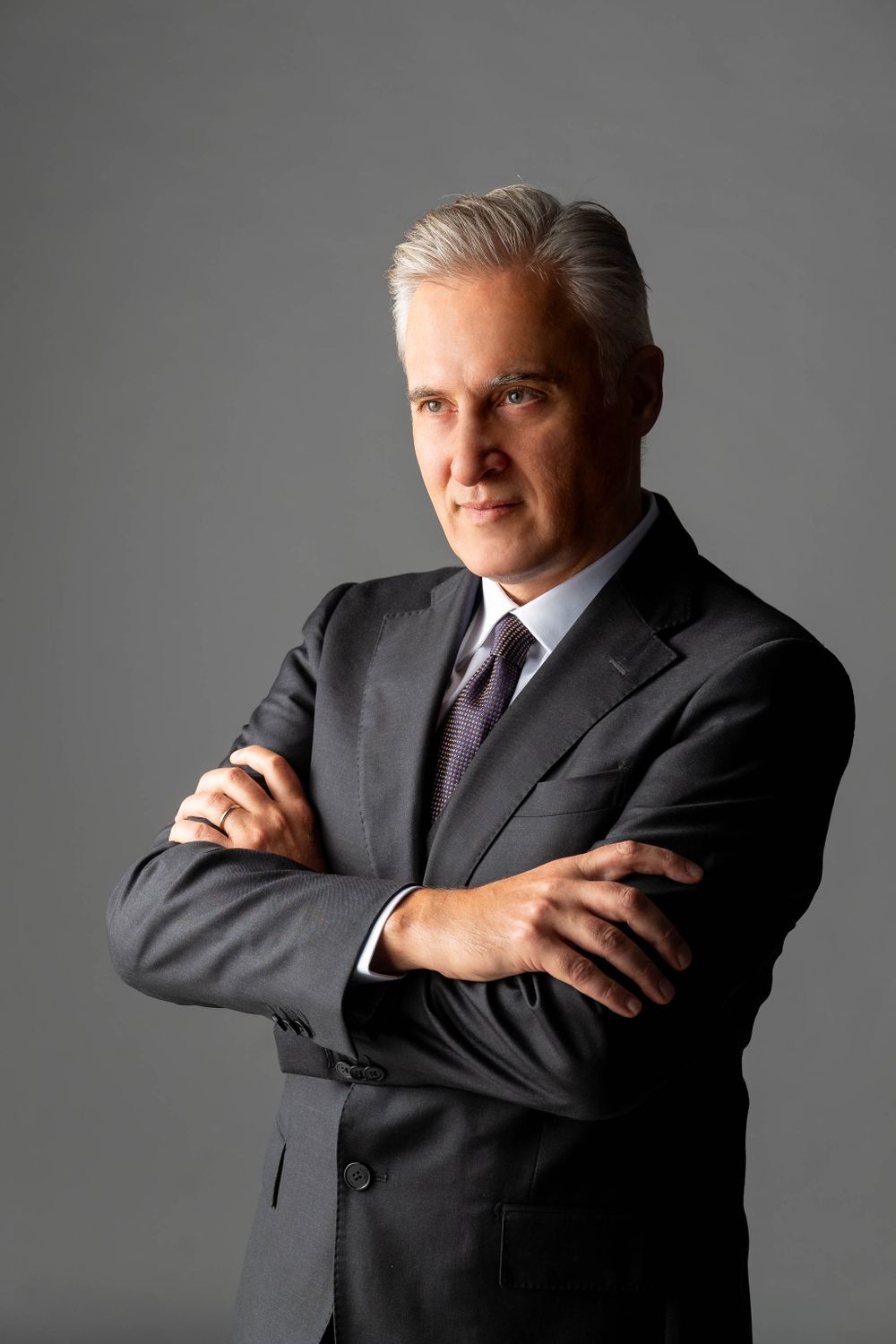 Professional portrait of a businessman in dark suit with silver hair against gray background.