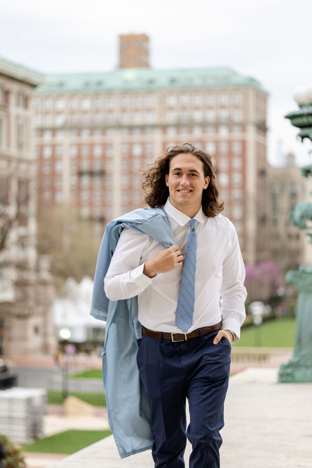 Graduate in professional attire holds Columbia graduation gown against campus backdrop.