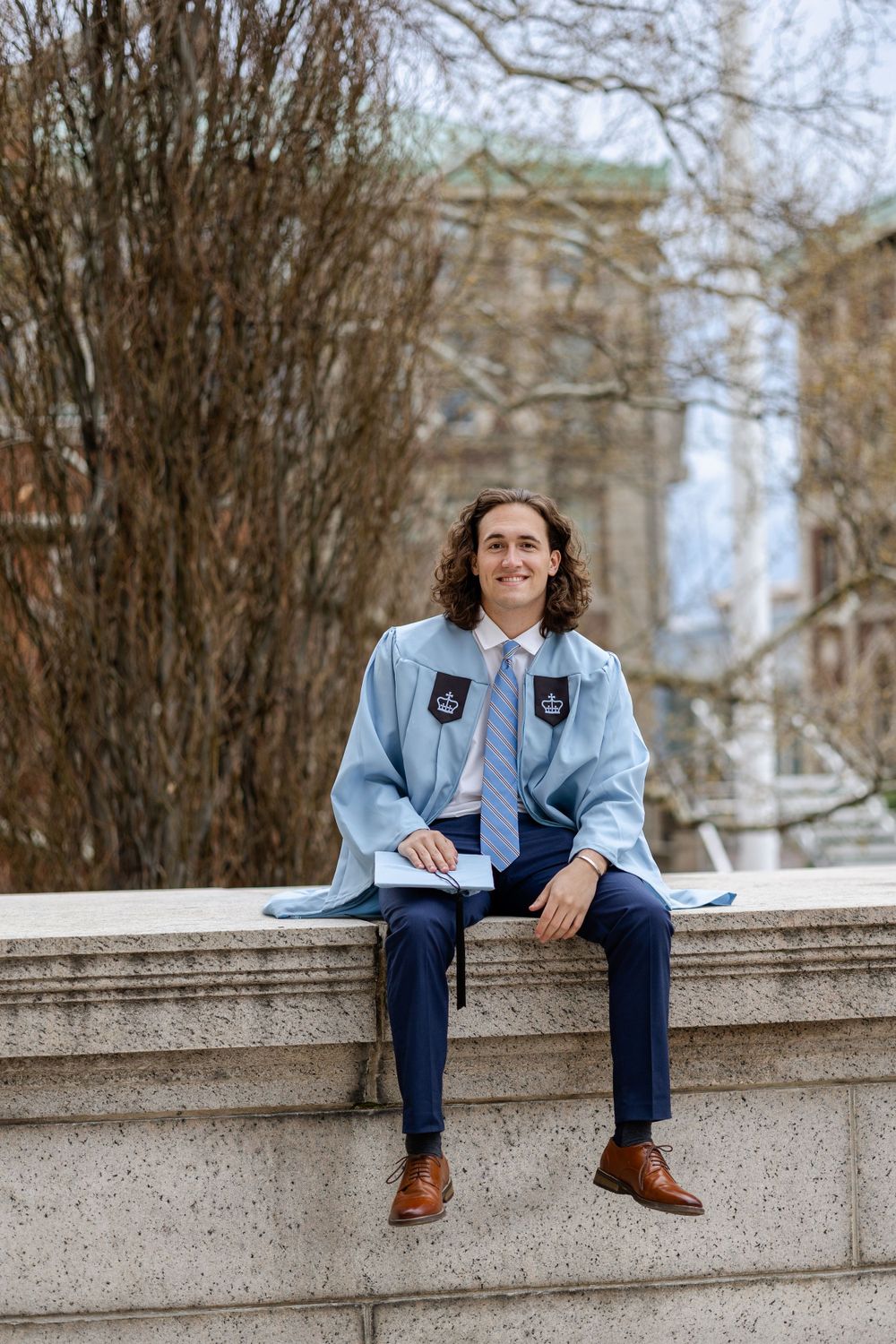 A student in graduation attire sits on a stone wall with campus buildings visible in the background.