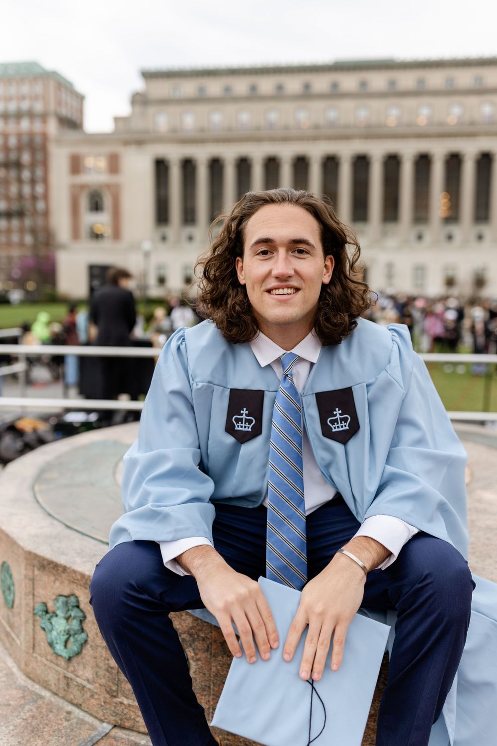 A graduate in light blue regalia sits on a stone ledge in front of Columbia University's iconic architecture.