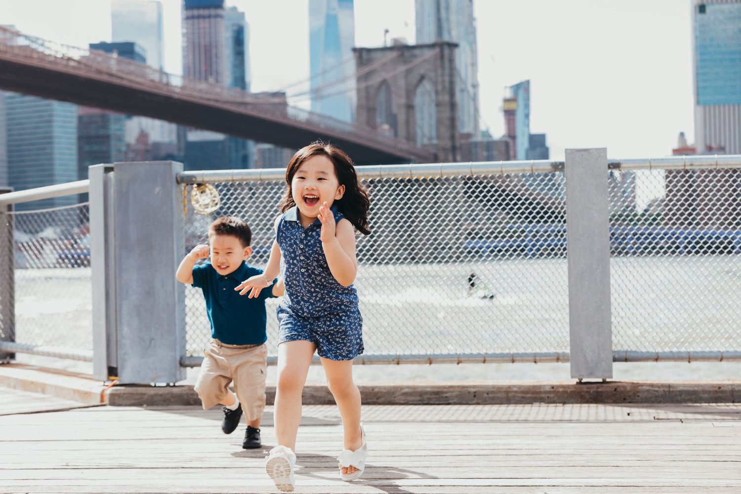 Two young Asian children running and laughing on a wooden boardwalk with the Brooklyn Bridge skyline in the background.