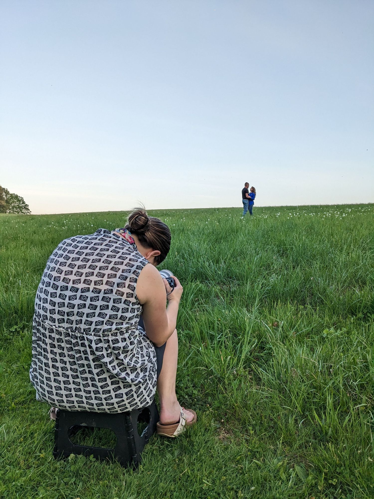 Photographer with long lens camera crouches in green field while subject stands in distance.