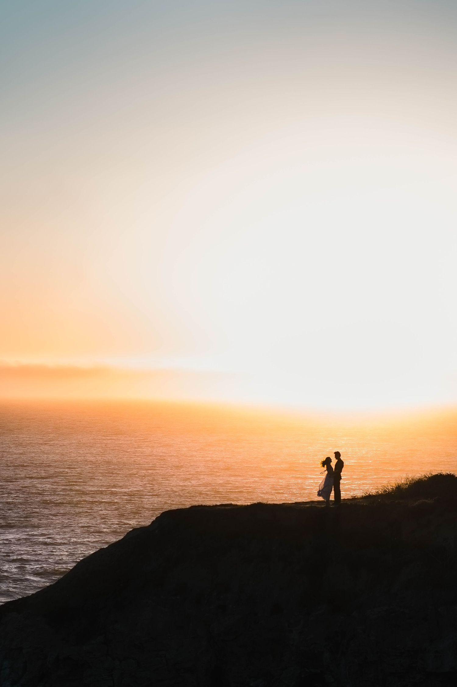 Silhouetted figures stand on a cliff edge against a dramatic orange sunset over the ocean.