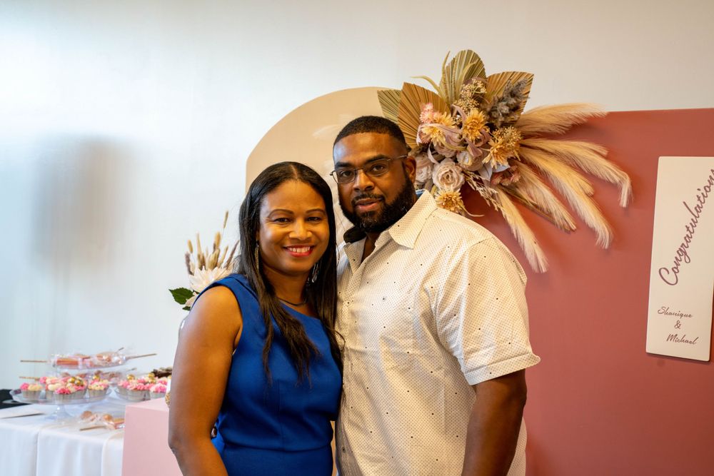 A couple in formal attire stands together at an event with a decorative floral arrangement in the background.