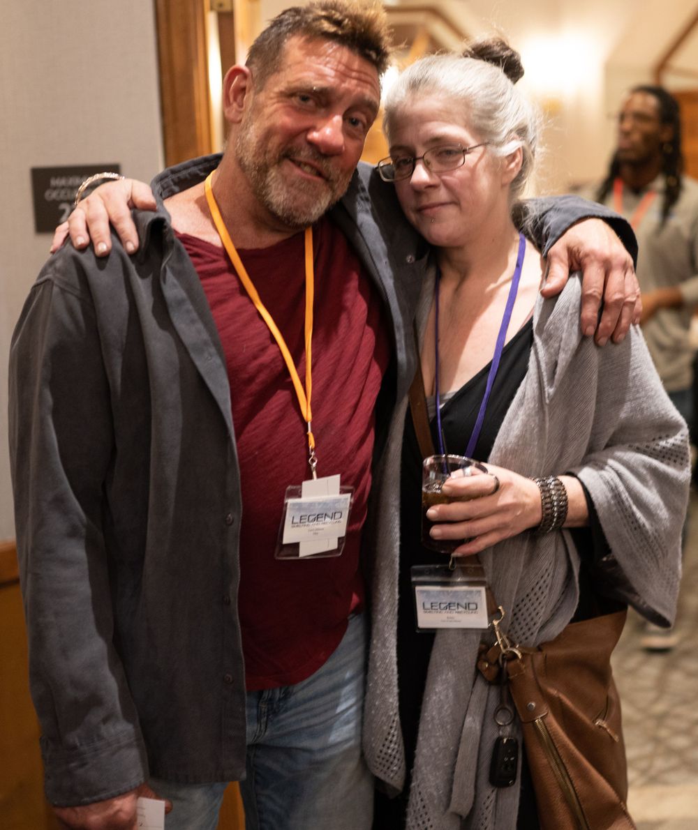 Two conference attendees wearing lanyards pose together at an indoor event, standing in a hallway.