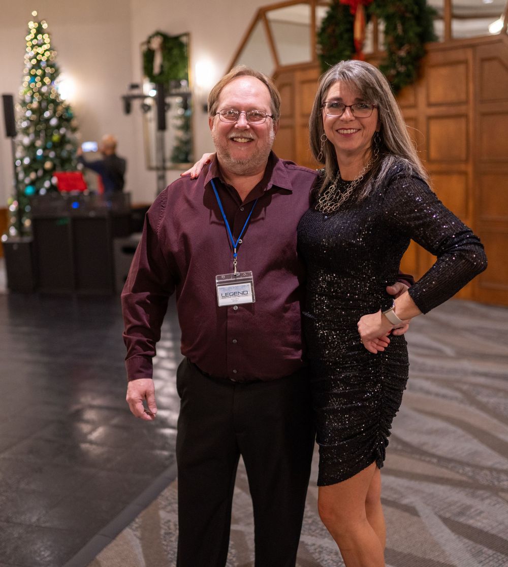A couple poses together at a holiday party with a decorated Christmas tree visible in the background.