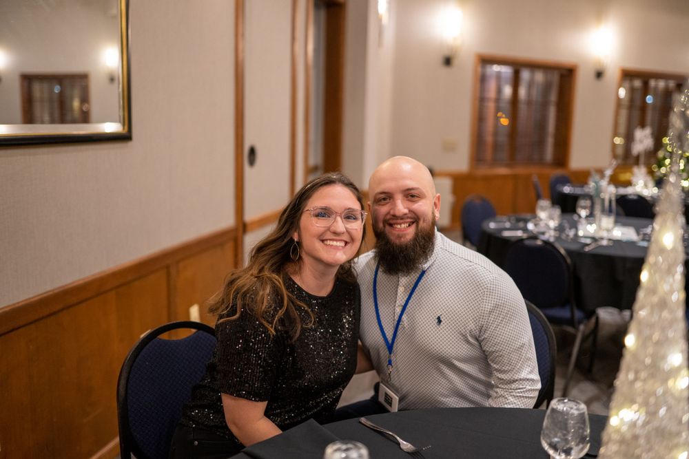 A couple smiles warmly while sitting together at an evening event with dining tables visible in the background.