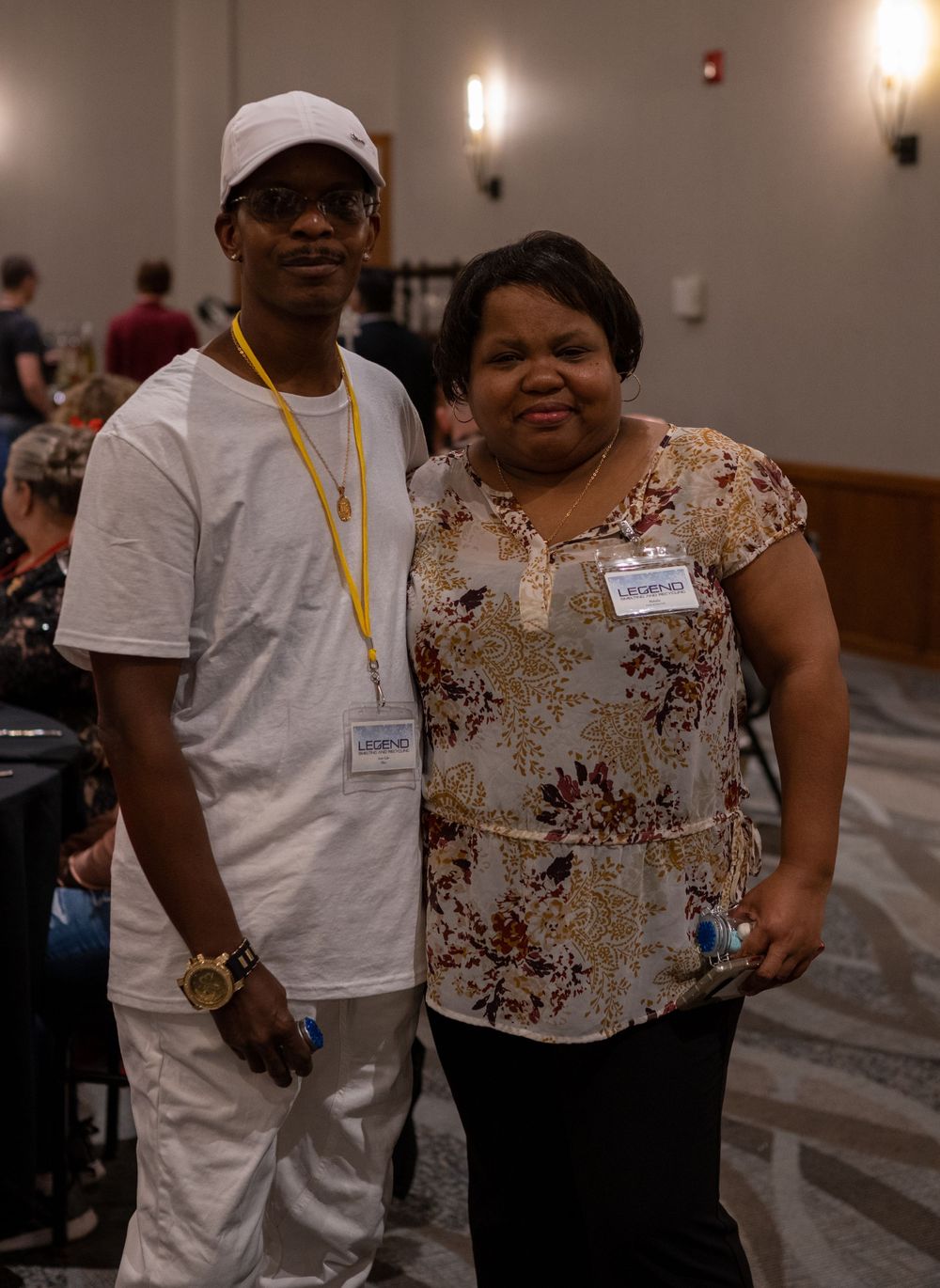 Two attendees wearing name tags pose together at an indoor event with a floral patterned shirt and white outfit.