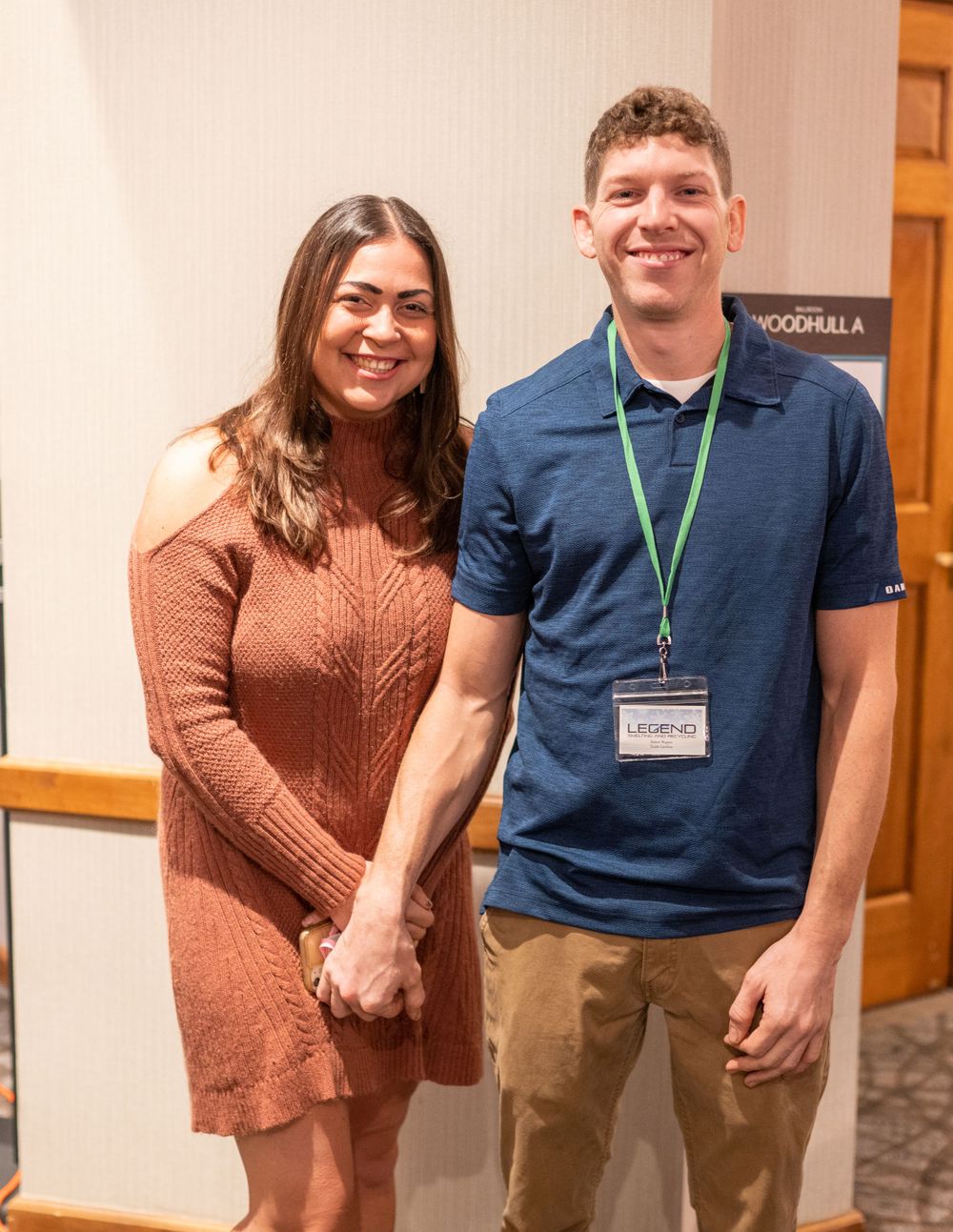 Two people in casual attire smile at the camera while standing together at an indoor event.