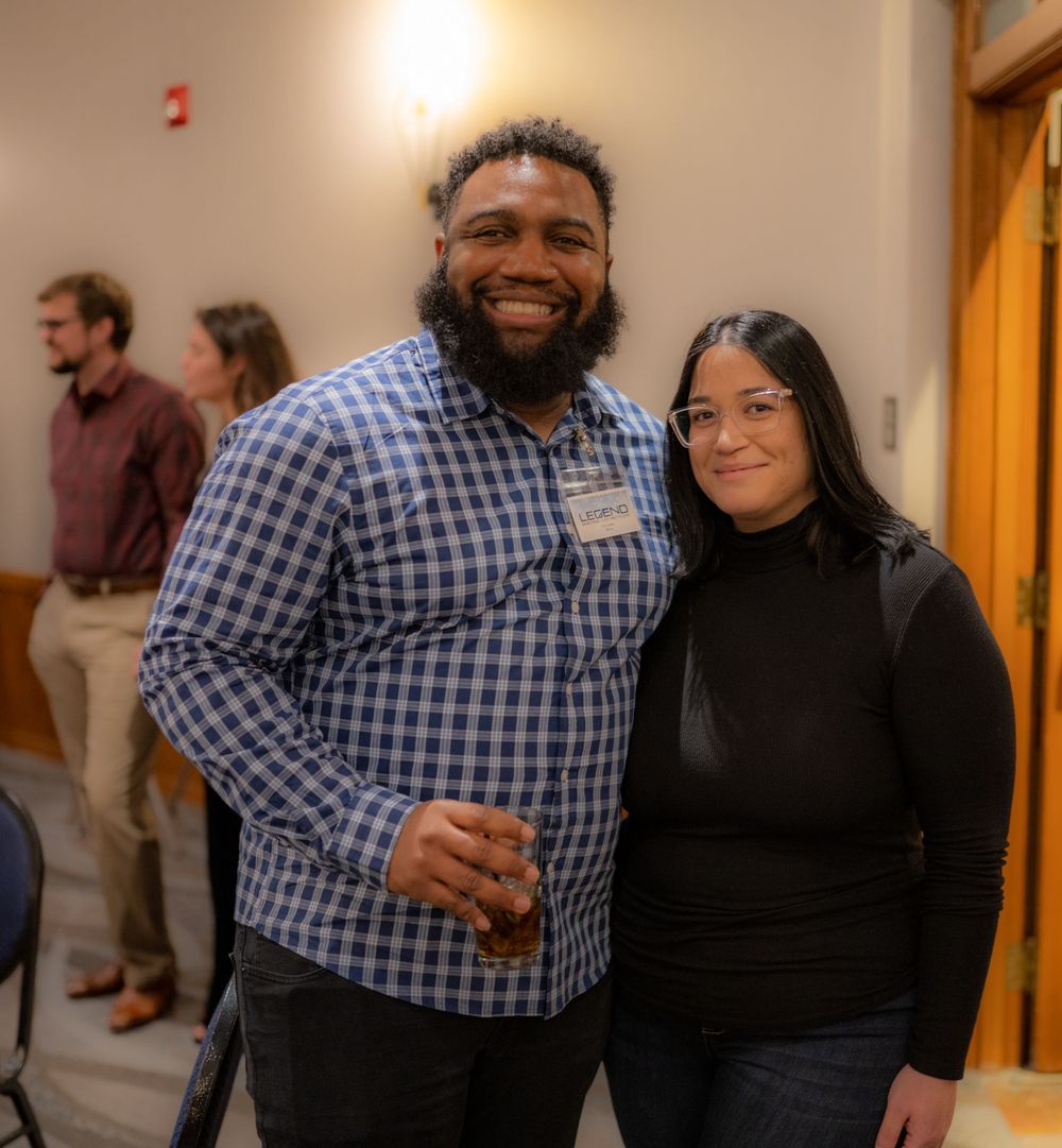Two people at an indoor social event stand close together smiling warmly at the camera.