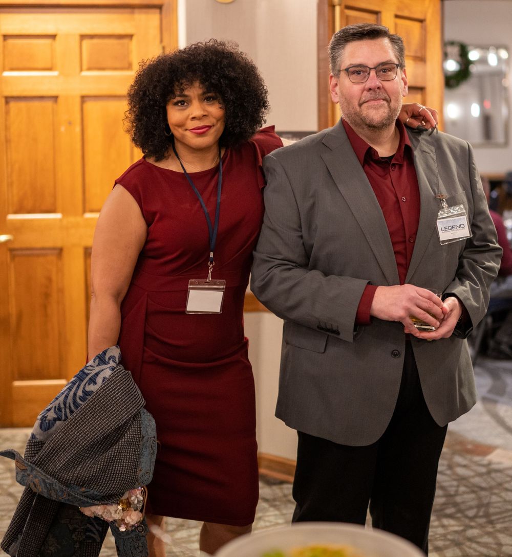Two professionals wearing name tags and burgundy attire smile together at a formal indoor event.