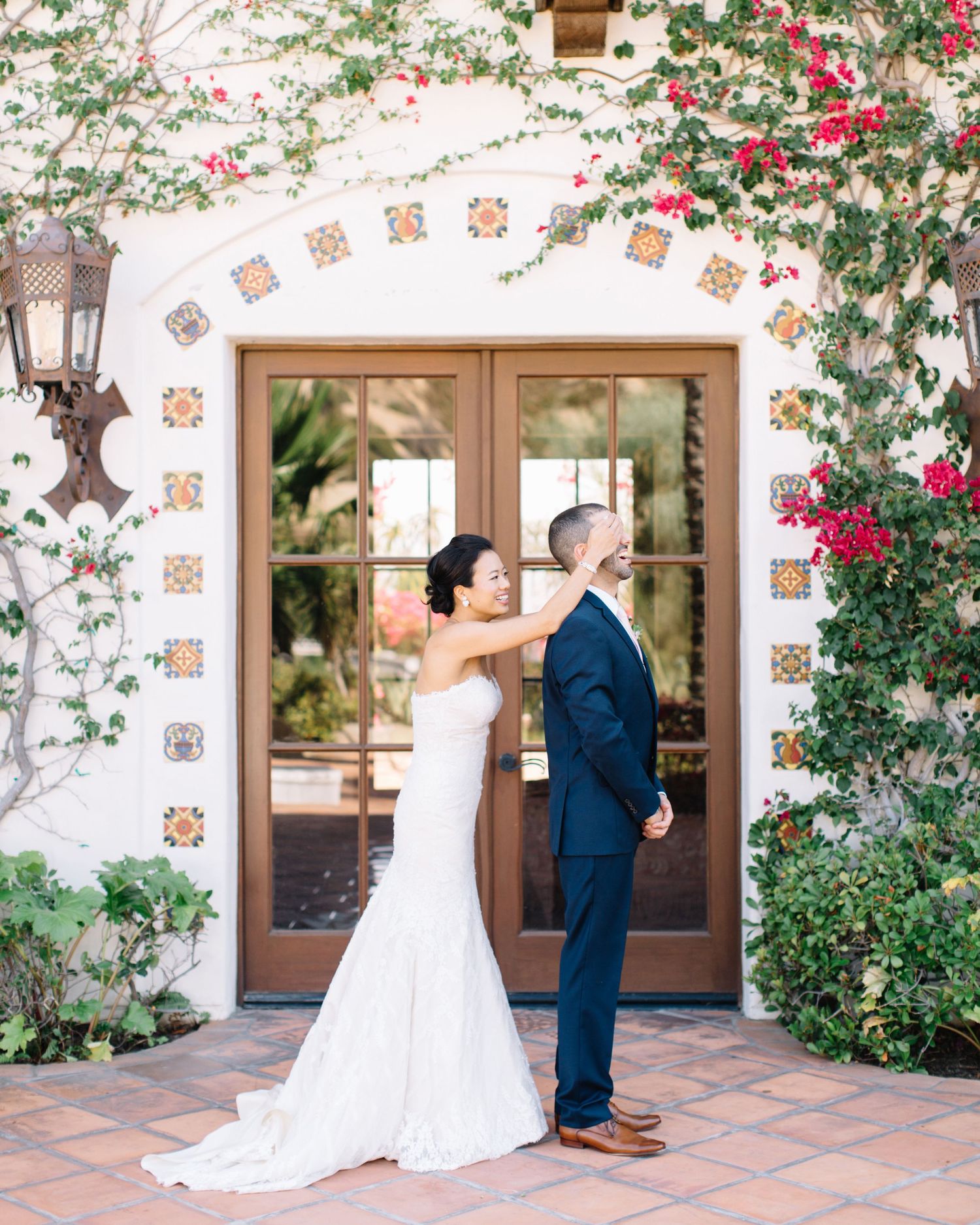 Playful fine art wedding photo of a bride covering her groom's eyes in front of a floral Spanish-style archway.
