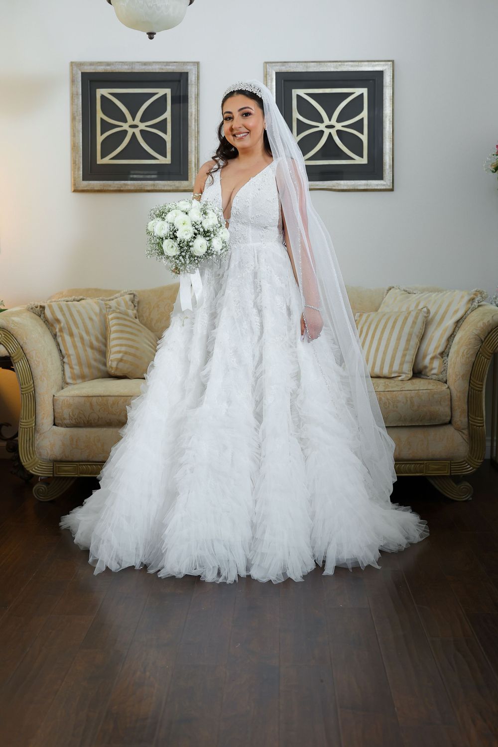 A stunning wedding dress with a ruffled tulle skirt, plunging neckline, and flowing veil stands out against dark hardwood floors.