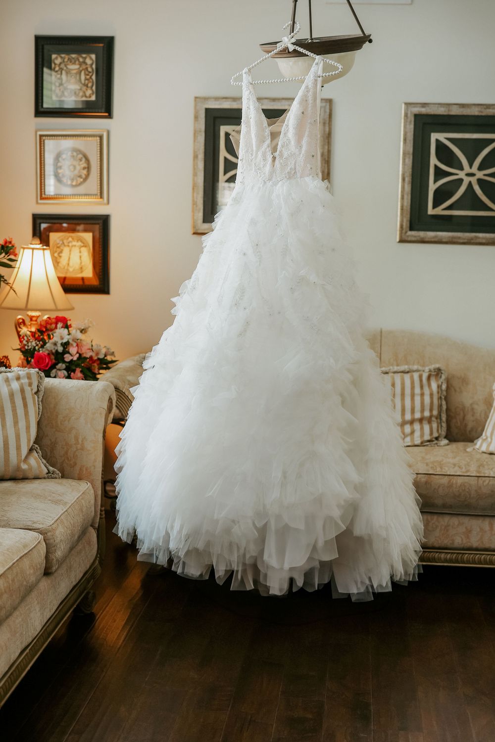 White wedding dress hanging in a cozy living room with hardwood floors and framed artwork on beige walls.