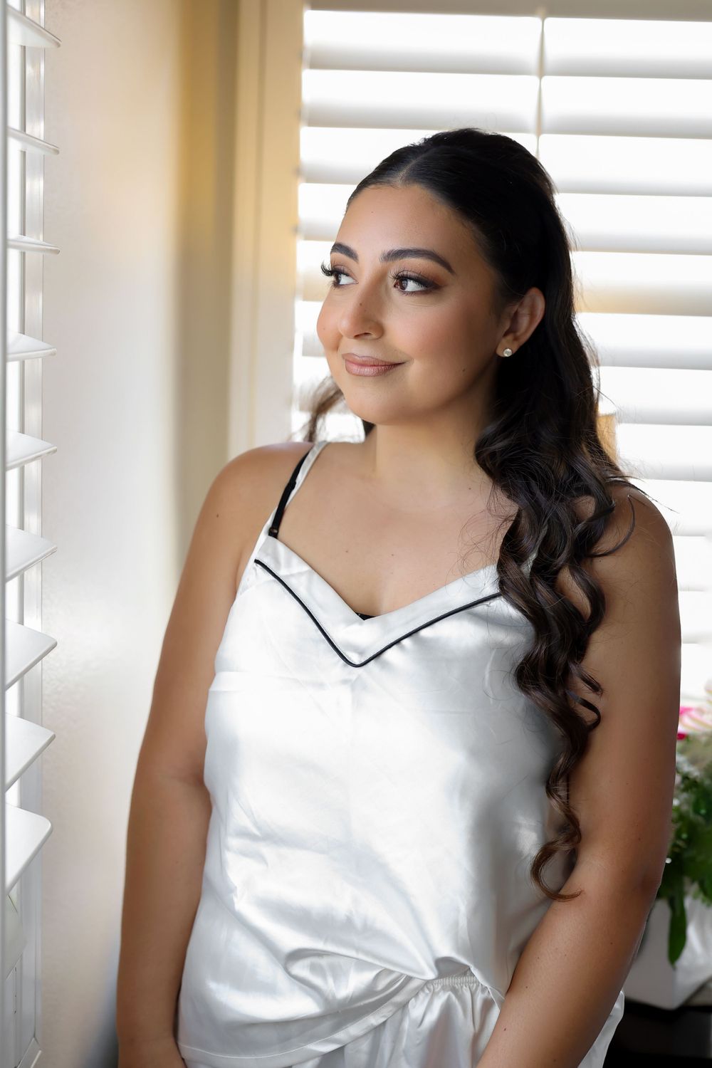 A person in a white satin camisole top poses in front of window blinds with a soft smile.