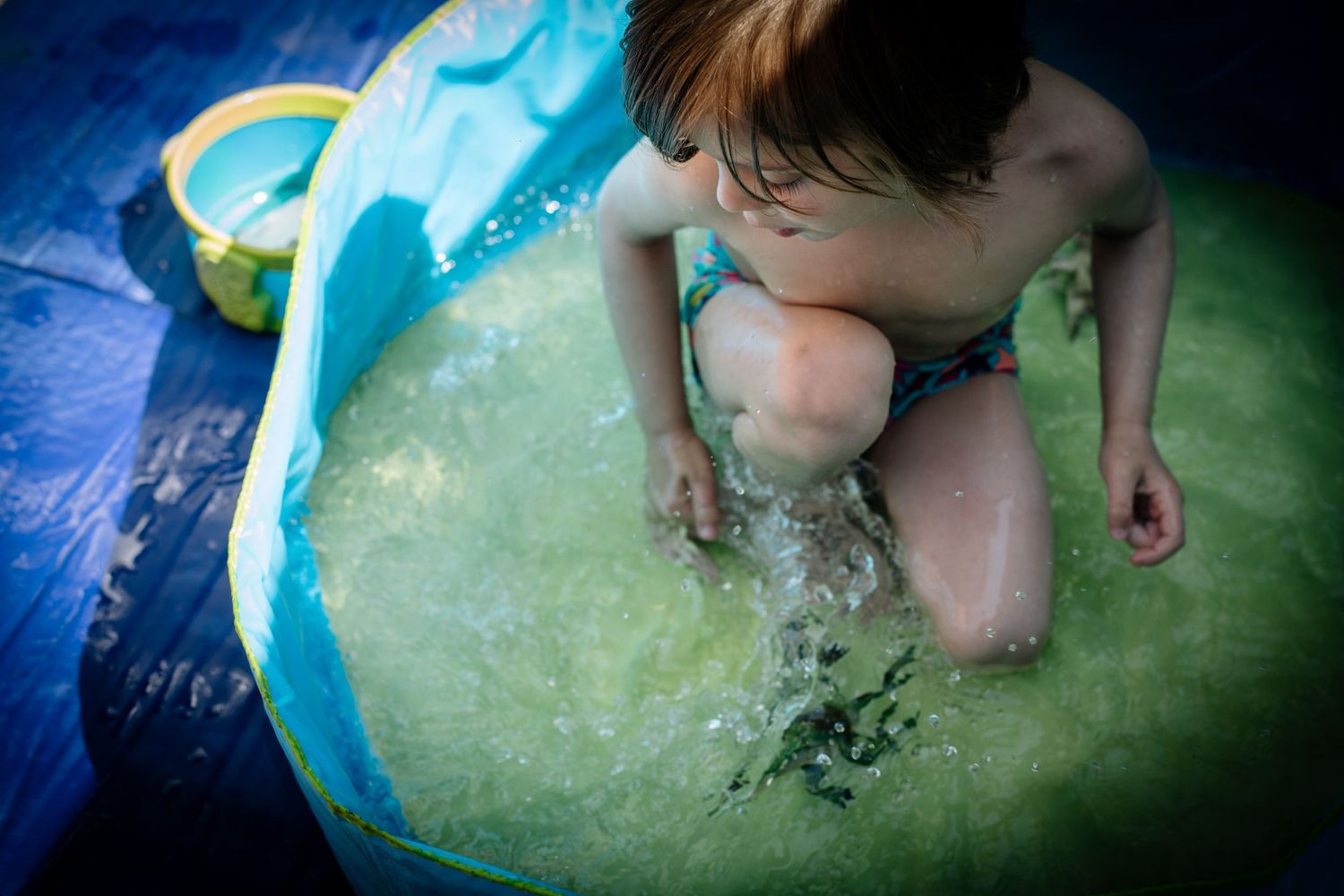 Small child playing in a blue and green inflatable pool with water.