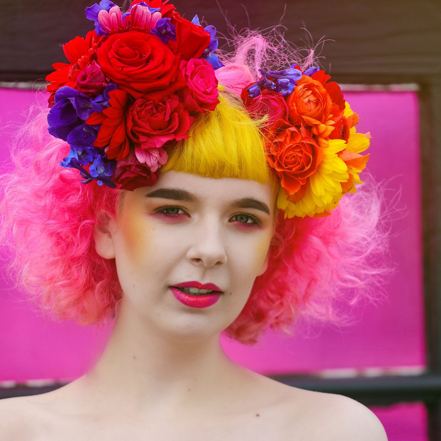 Pink, yellow haired model with brightly coloured makeup and rainbow floral head buns smiling with pink graffiti behind.