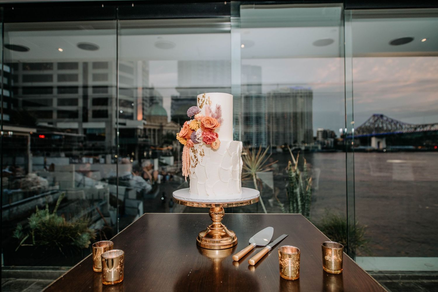 Elegant white wedding cake with coral flowers displayed on gold stand with candlelit reception backdrop.