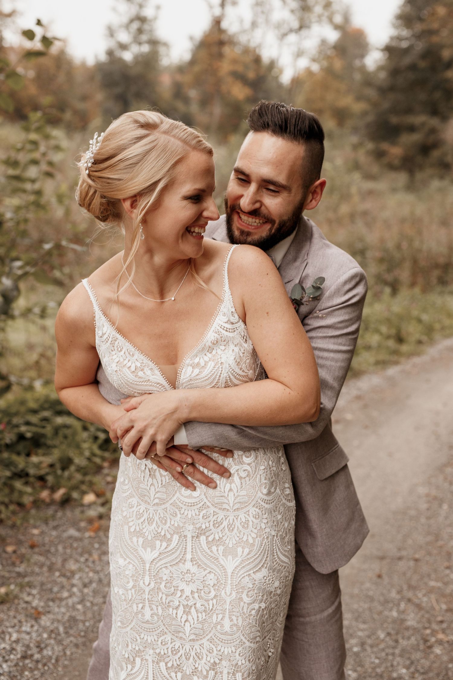 Ein romantisches Hochzeitspaar umarmt sich und lacht gemeinsam auf einem Kiesweg, umgeben von Herbstlaub.