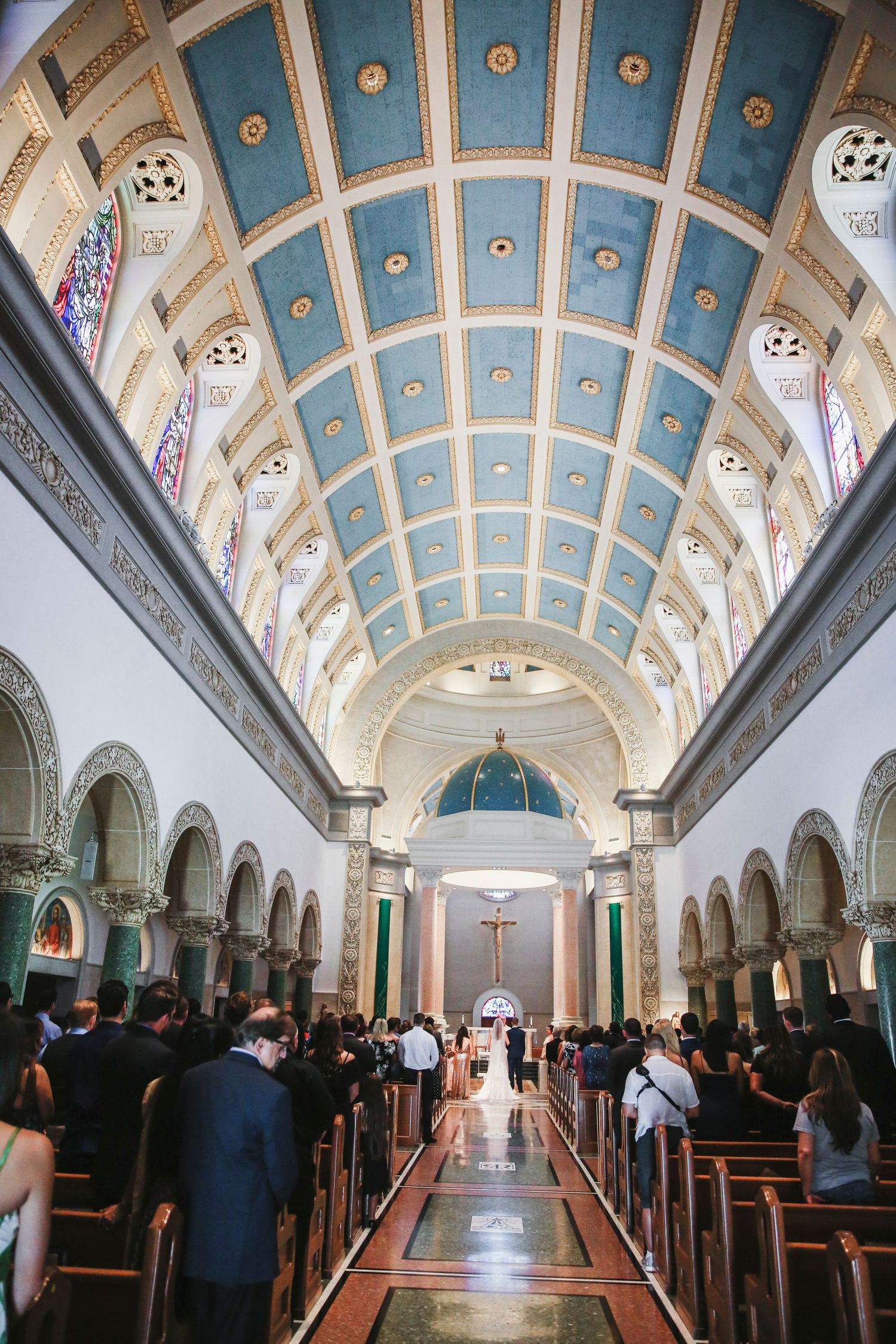 Interior view of a church with blue vaulted ceiling, arched columns and stained glass windows during a service.