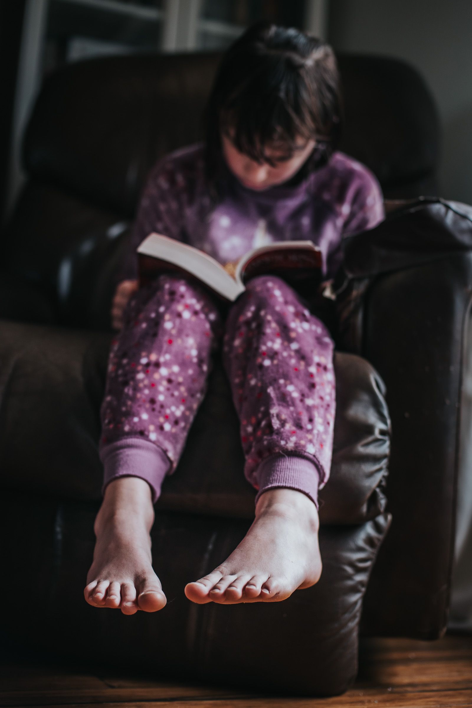 Focus on the feet of a girl sitting in a chair in purple pyjamas reading Warrior Cats.