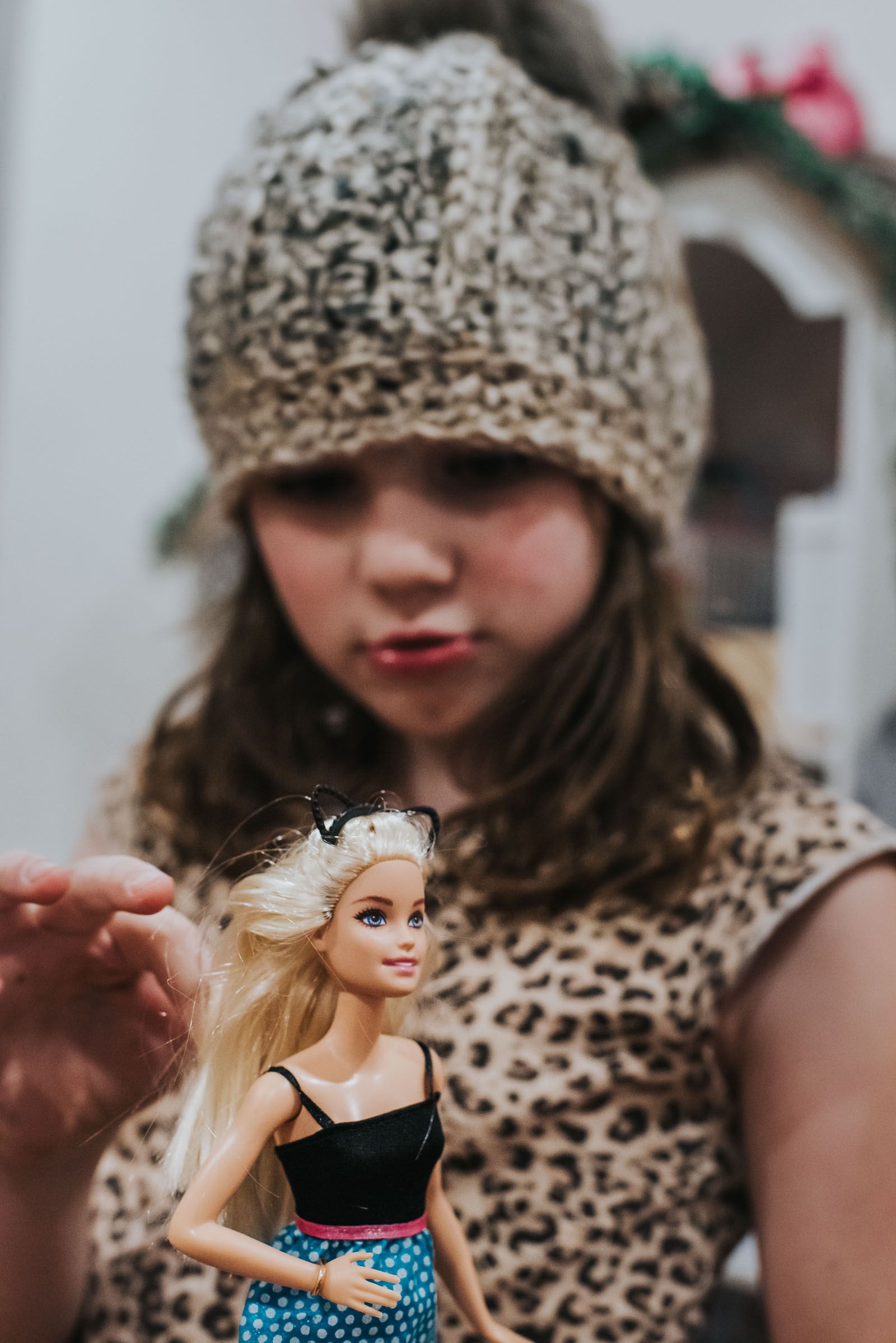 A girl is out of focus in a wool tuque, she's holding a Barbie doll with a cat headband.