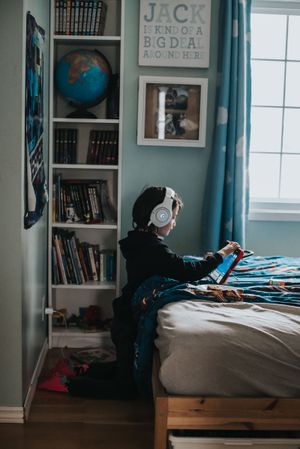 A young boy kneels facing a bed, he's wearing white headphones and playing on a tablet.