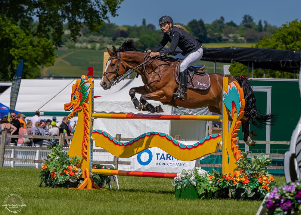 Horse and rider jumping over decorated turquoise and orange obstacle during competition.