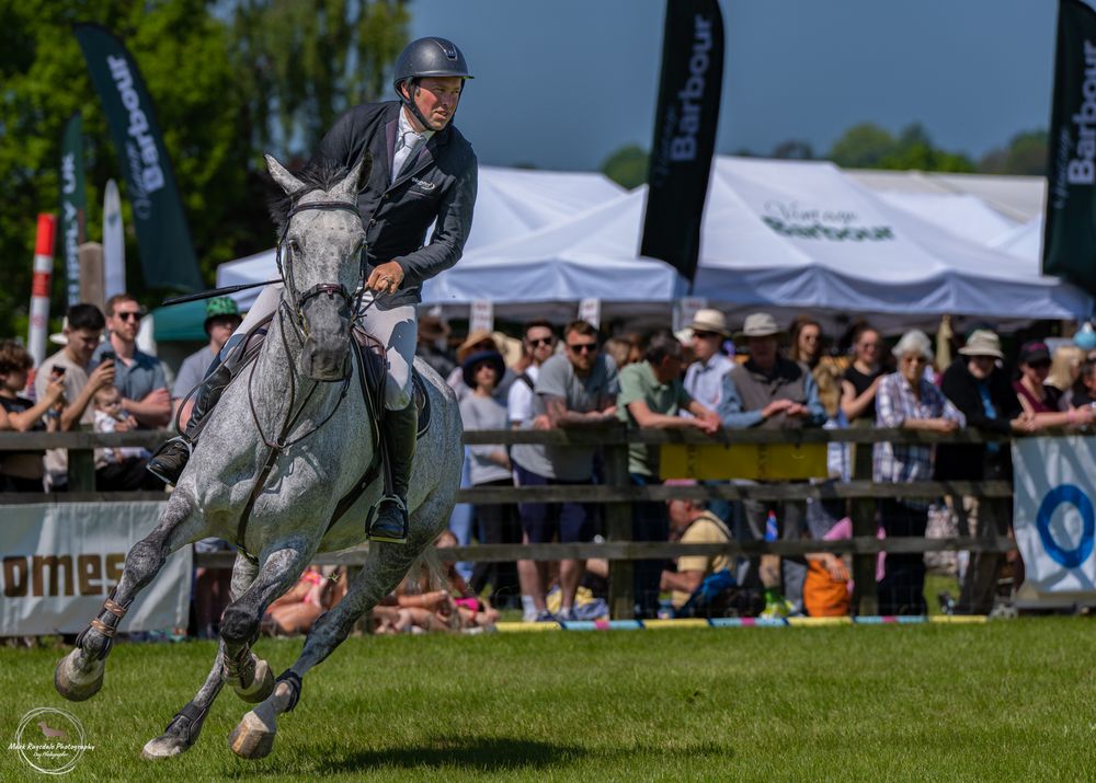 Grey horse and rider soaring over jump at outdoor equestrian event with spectators watching.