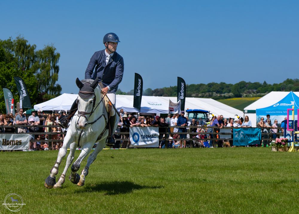 Horse and rider performing in outdoor showjumping event with spectators.