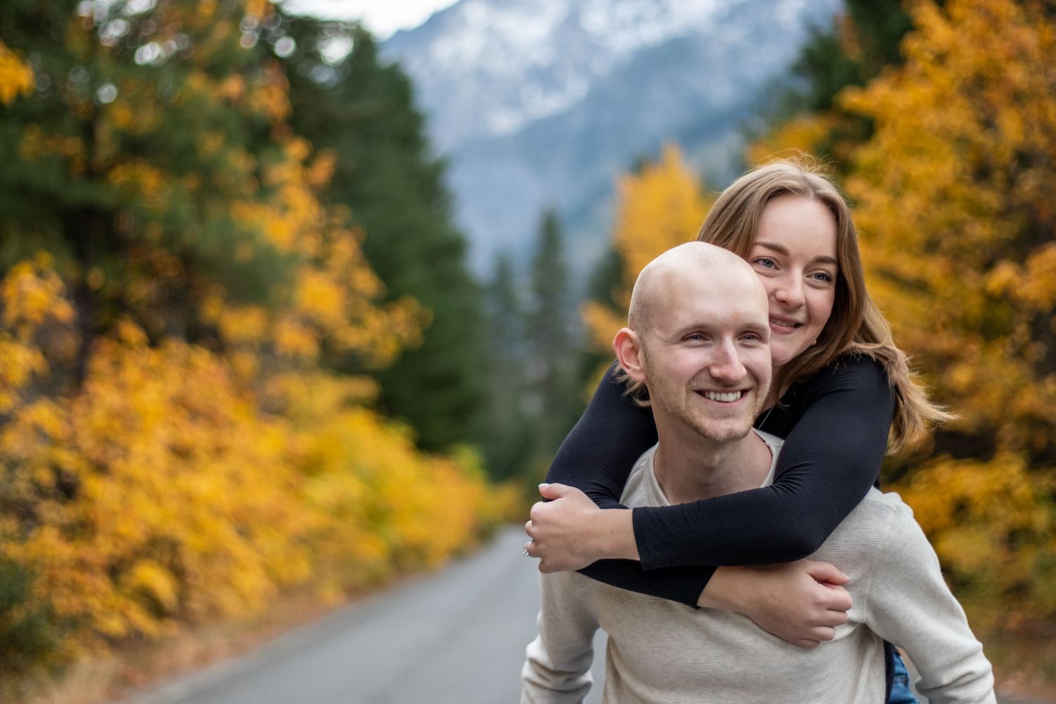 A playful piggyback ride moment on a mountain road lined with autumn trees.