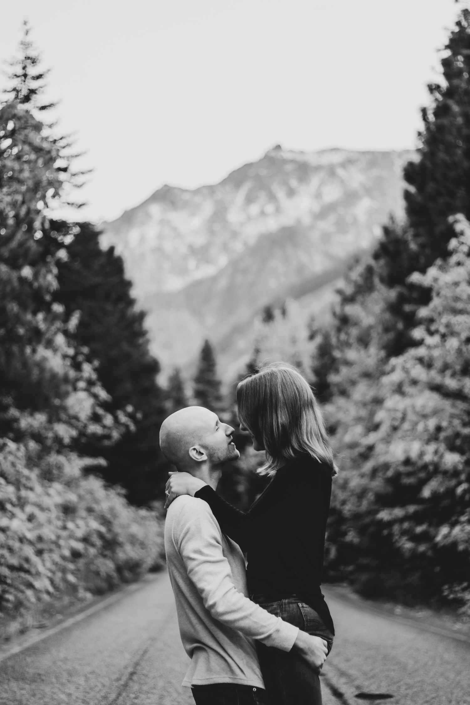 A black and white portrait of a couple embracing on a mountain road surrounded by trees.