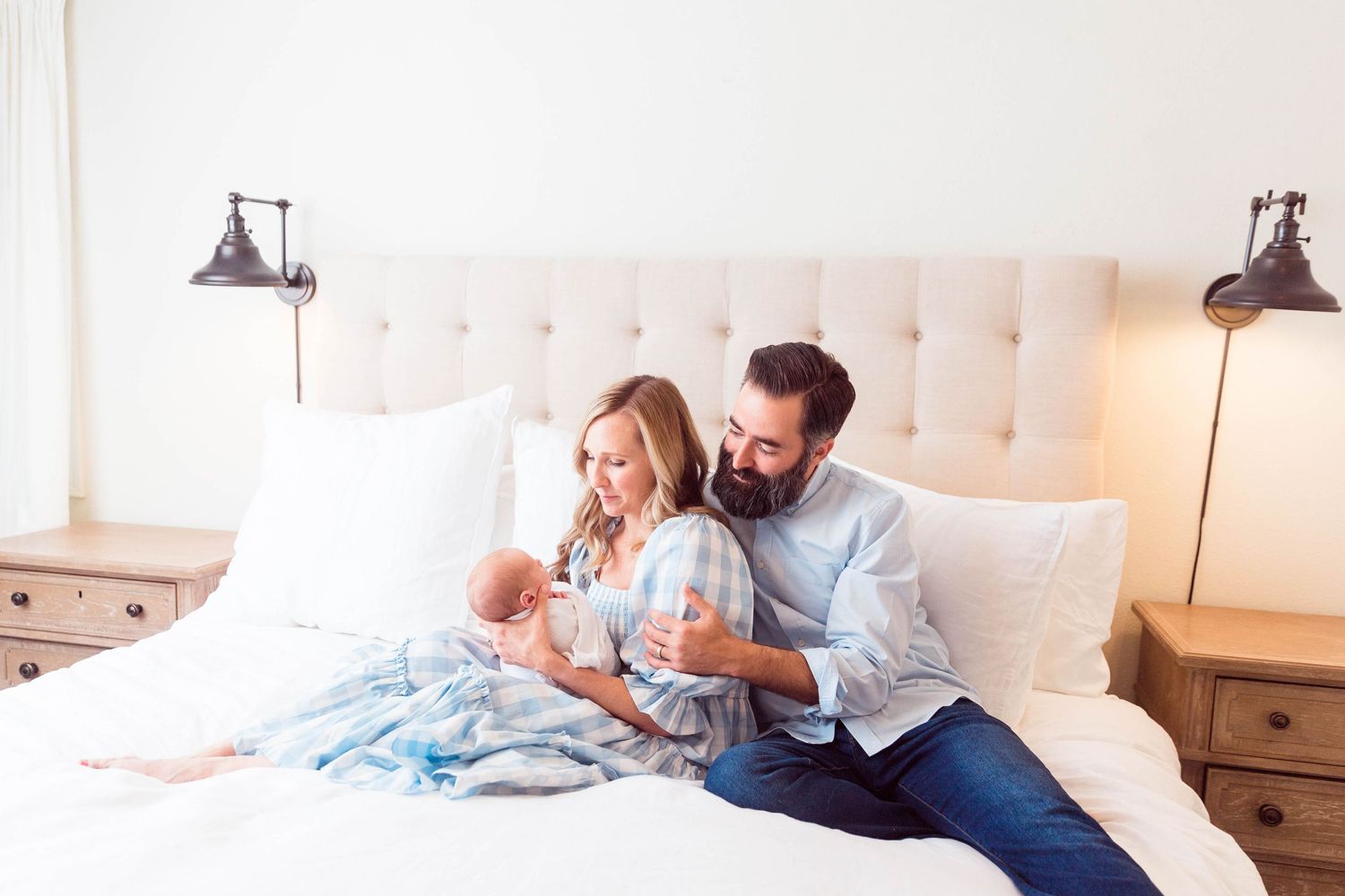 A couple in light blue and white attire cuddle with their newborn baby on a beige tufted headboard bed.