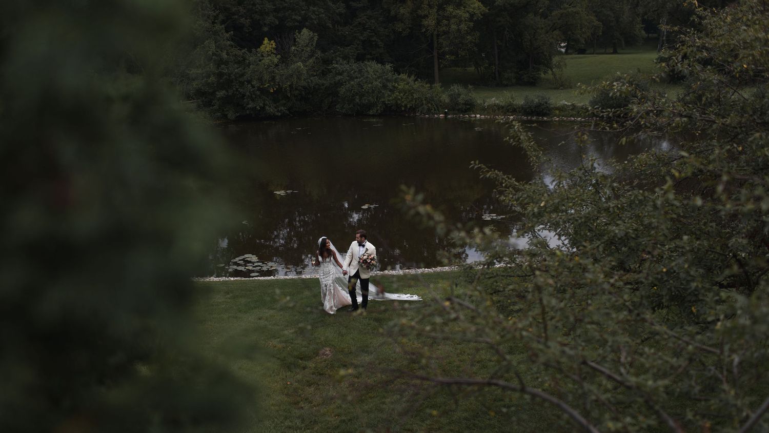 Two figures surrounded by lush greenery sit together at a small table by a tranquil pond.