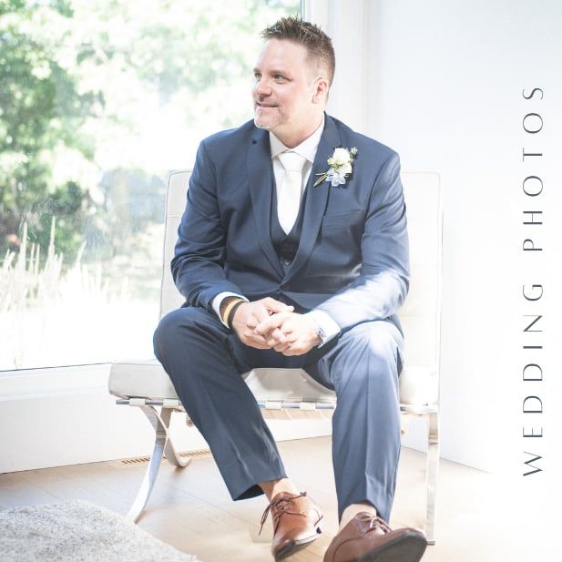 A groom in navy suit sits relaxed on a white bench in natural lighting.