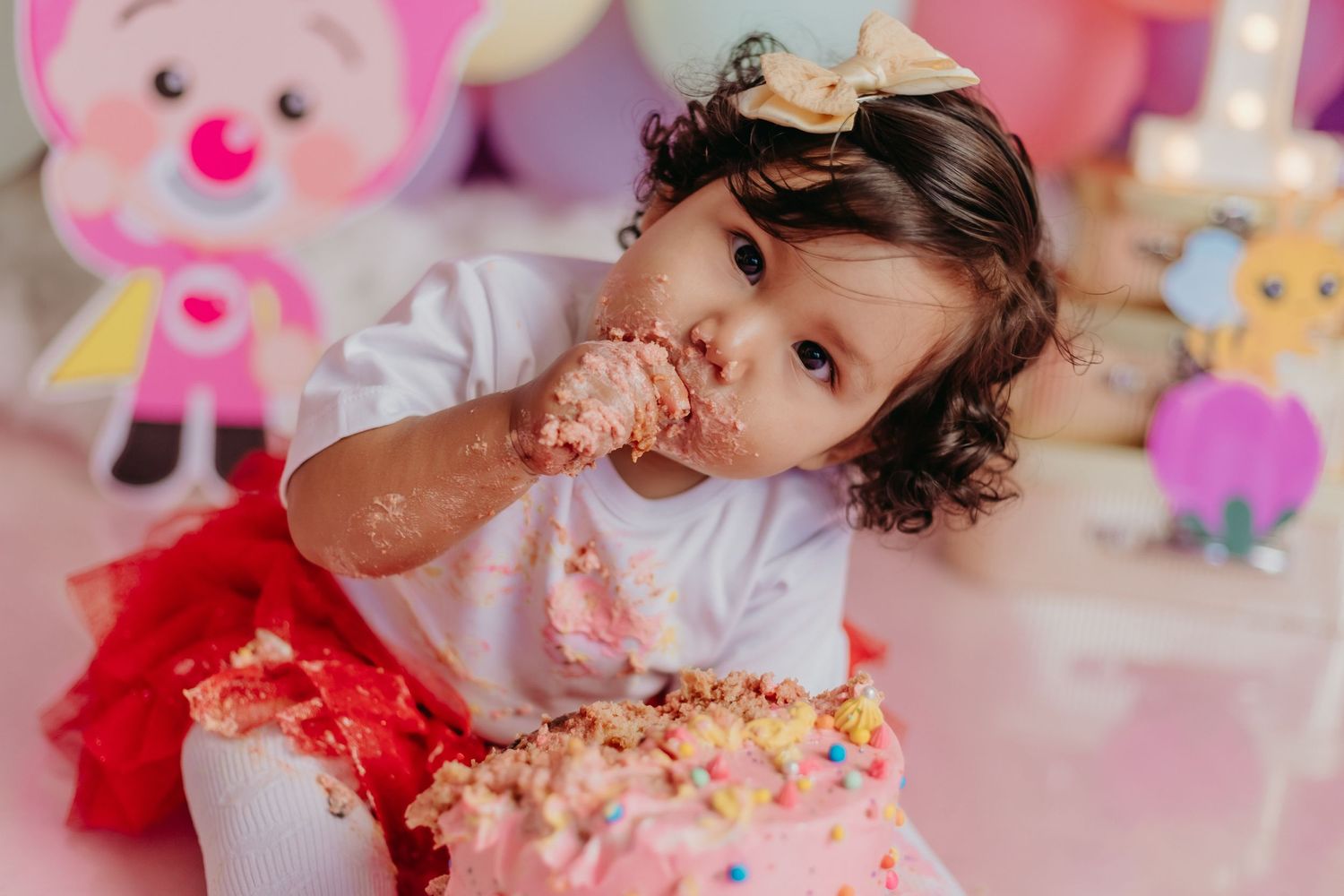 A toddler wearing a red tutu enjoys a frosted cake at a festive party with stuffed toys in the background.