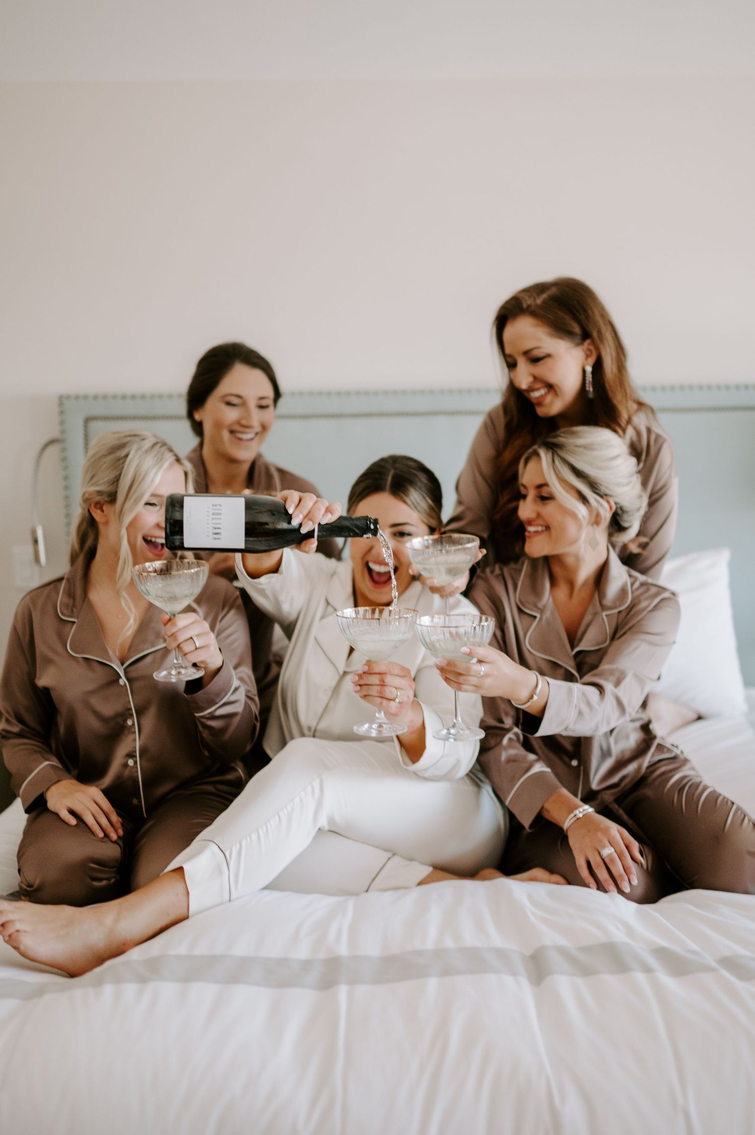 Friends in matching brown pajamas laugh together while relaxing on a bed.