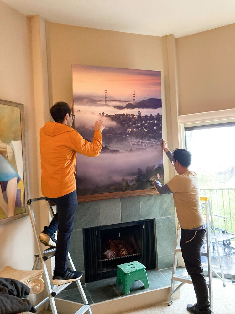 Living room interior with people installing large Golden Gate Bridge fog artwork above fireplace.
