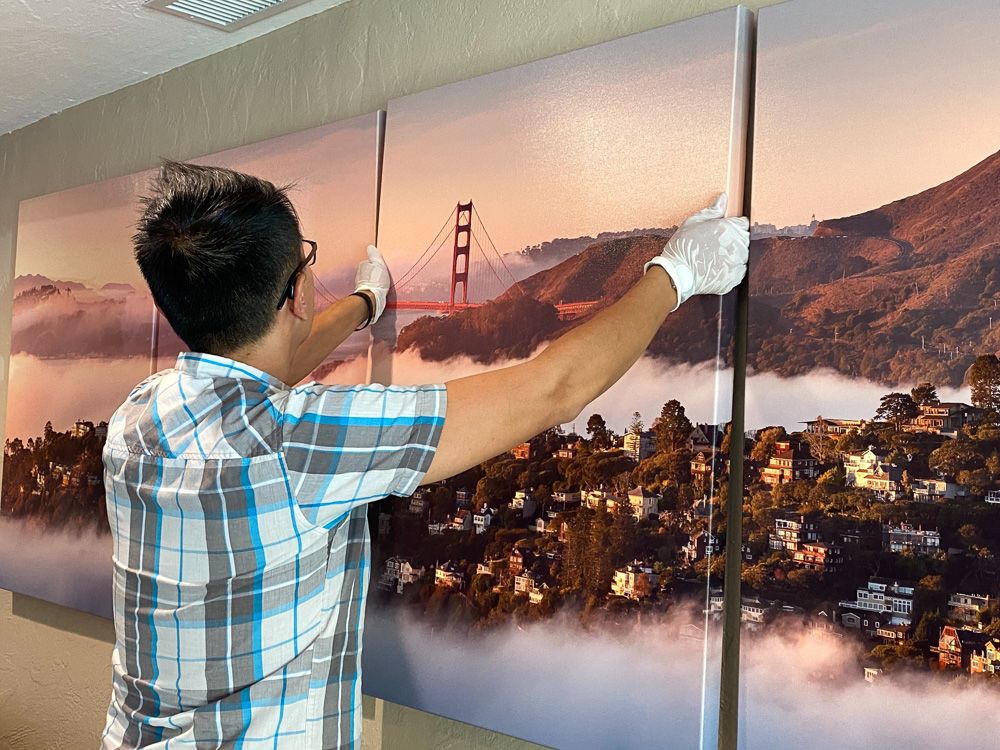 Person hanging a three-panel canvas print of San Francisco and Golden Gate Bridge on wall.