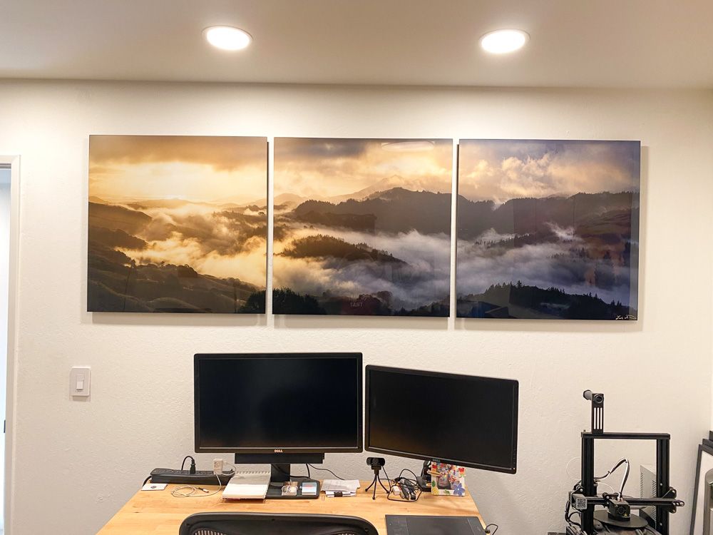 Three-panel canvas print of foggy mountain landscape mounted above dual monitor computer desk setup.