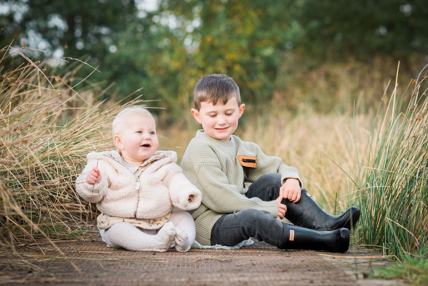 Little boy and baby girl sitting on a wooden bridge amongst grasses