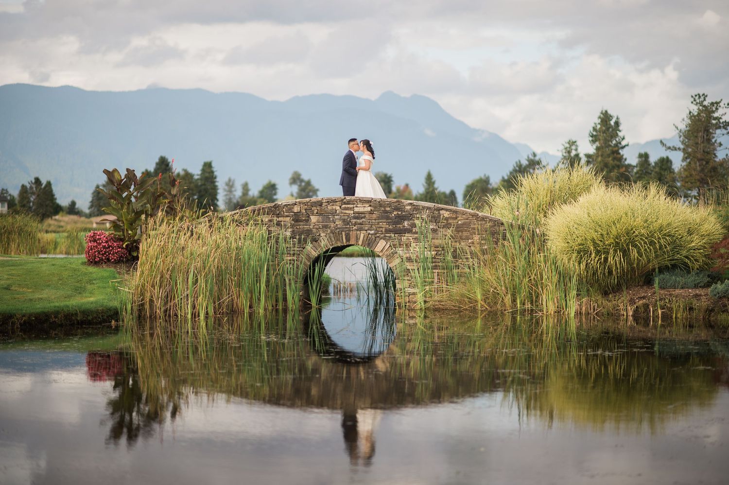 A couple stands on a stone bridge reflecting in still water with mountains in the background.