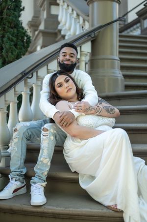 A romantic couple poses on elegant stone stairs, with the bride in a white gown and groom in casual wear.